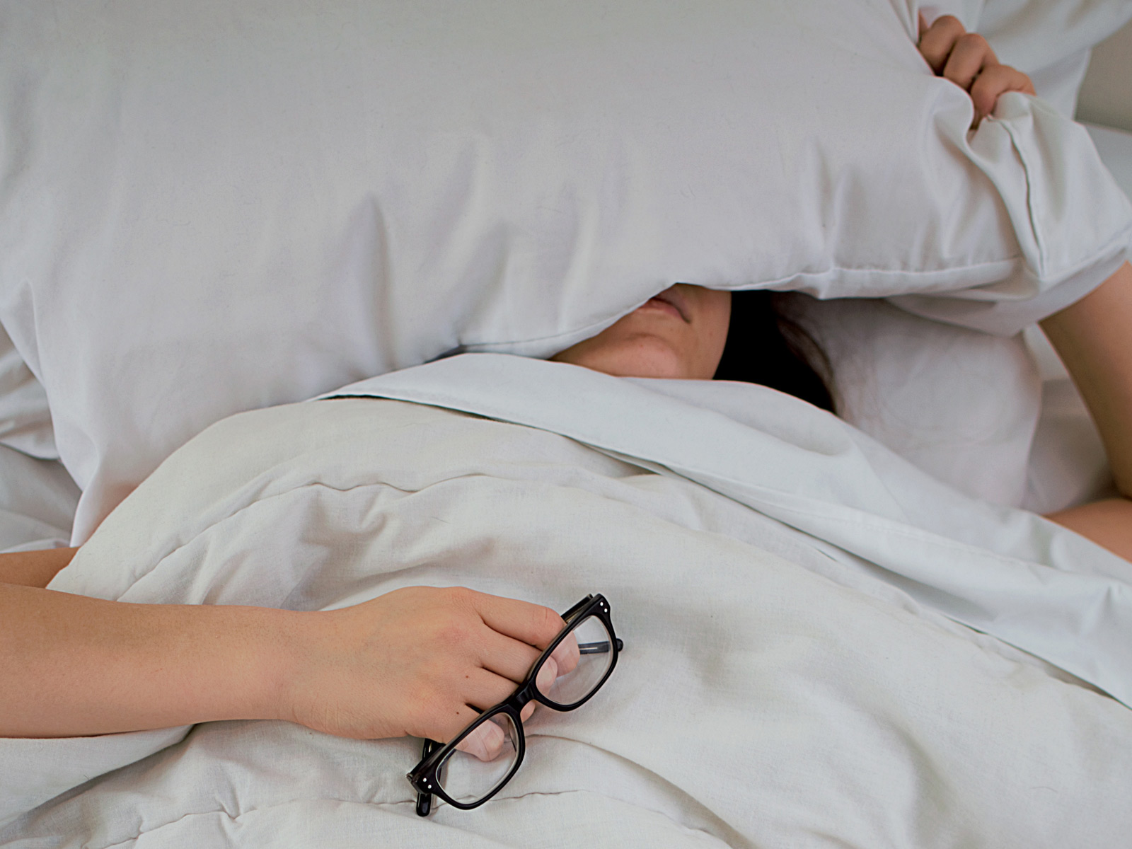 Woman with her head under the pillow with glasses in hand
